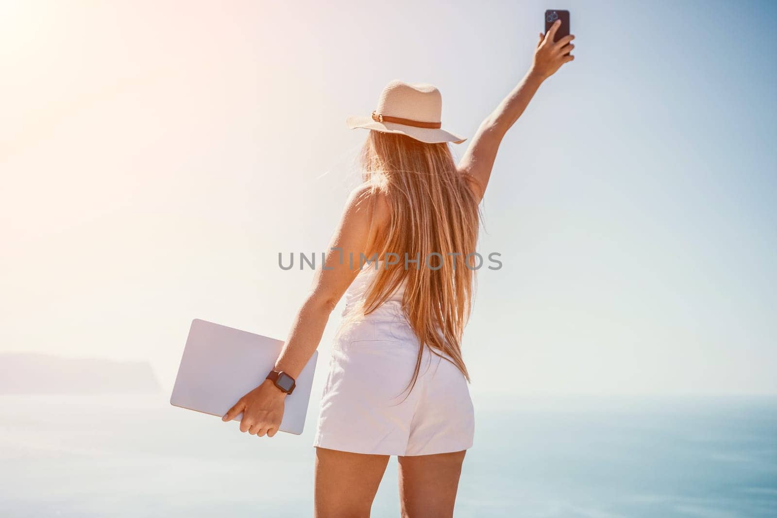 Successful business woman in yellow hat working on laptop by the sea. Pretty lady typing on computer at summer day outdoors. Freelance, travel and holidays concept.