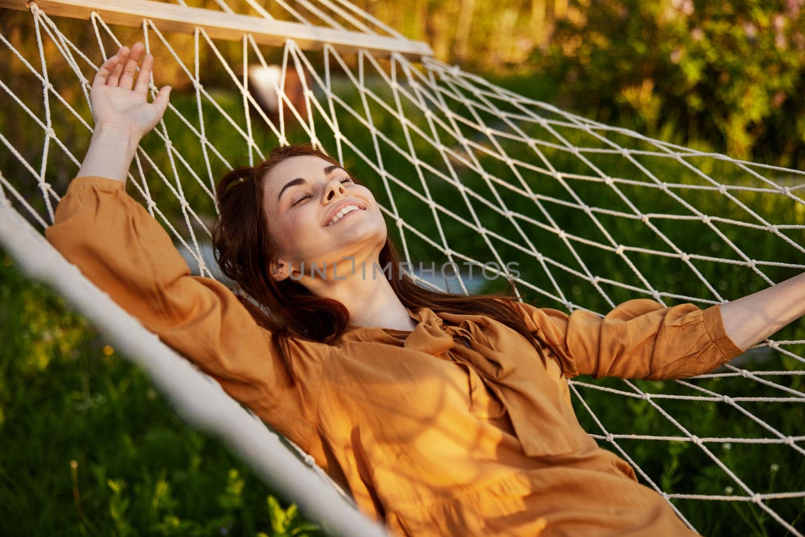 a happy woman is resting in a hammock with her eyes closed and her hands behind her head smiling happily enjoying the day by Vichizh