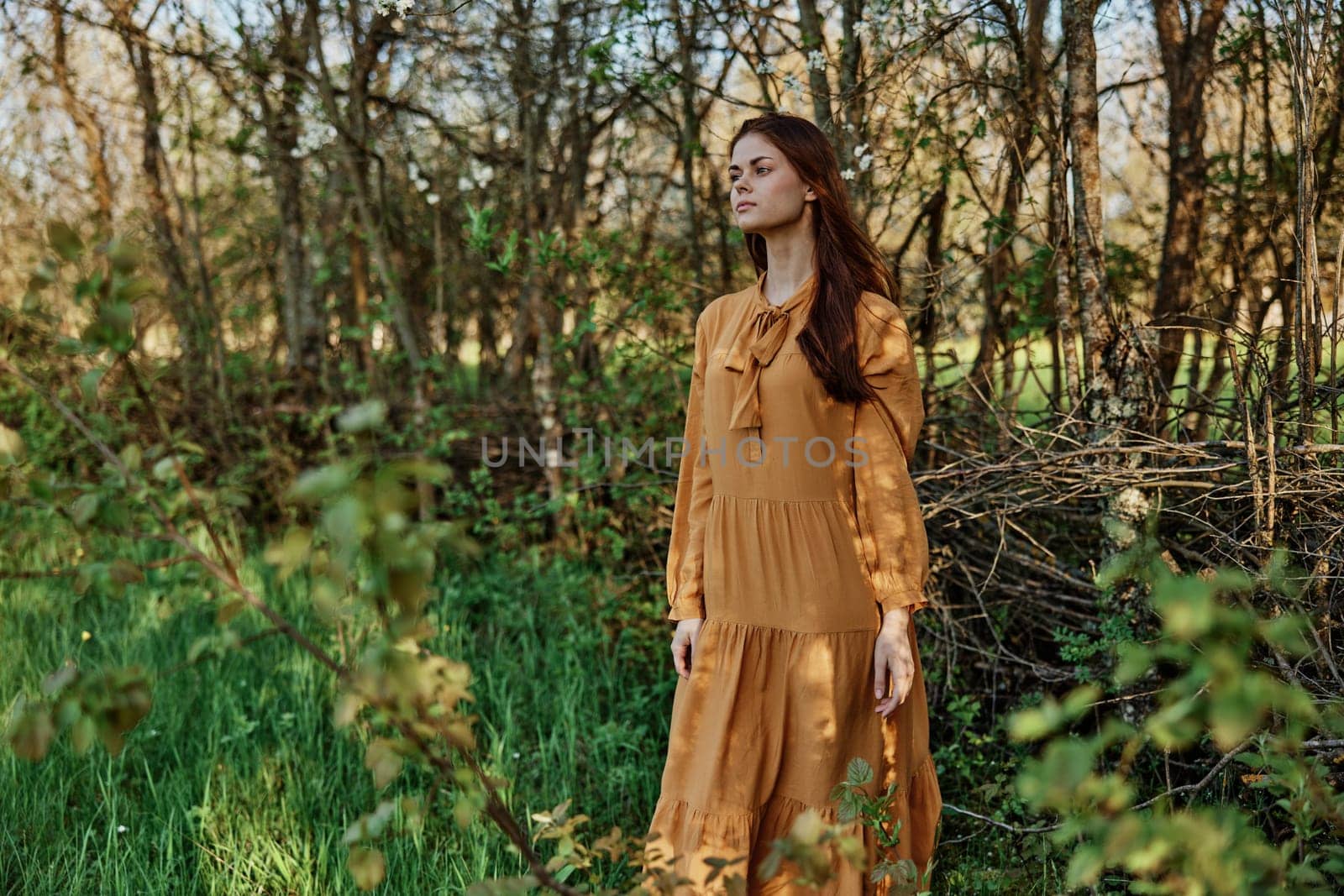 a woman with long hair walks in the shade near the trees, dressed in a long orange dress, enjoying the weather and the weekend, inspecting the trees. The theme of privacy with nature by Vichizh