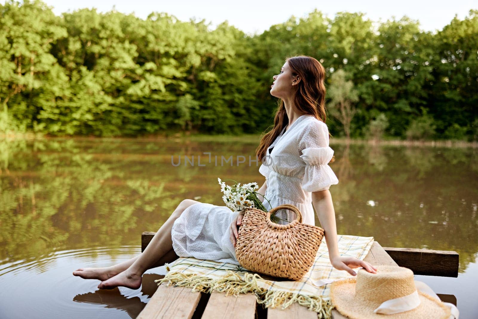 an elegant woman in a light dress sits on a pier by the lake with her feet in the water by Vichizh