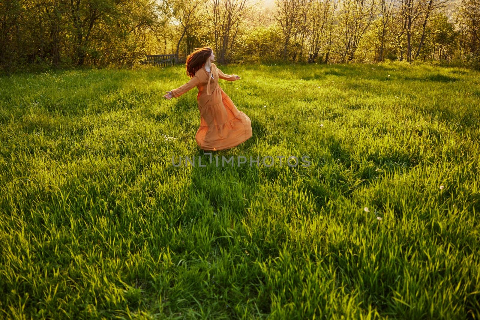 an attractive, slender, red-haired woman stands far away in a wide, green field during sunset in a long orange dress enjoying unity with nature and relaxation standing with her back to the camera. High quality photo