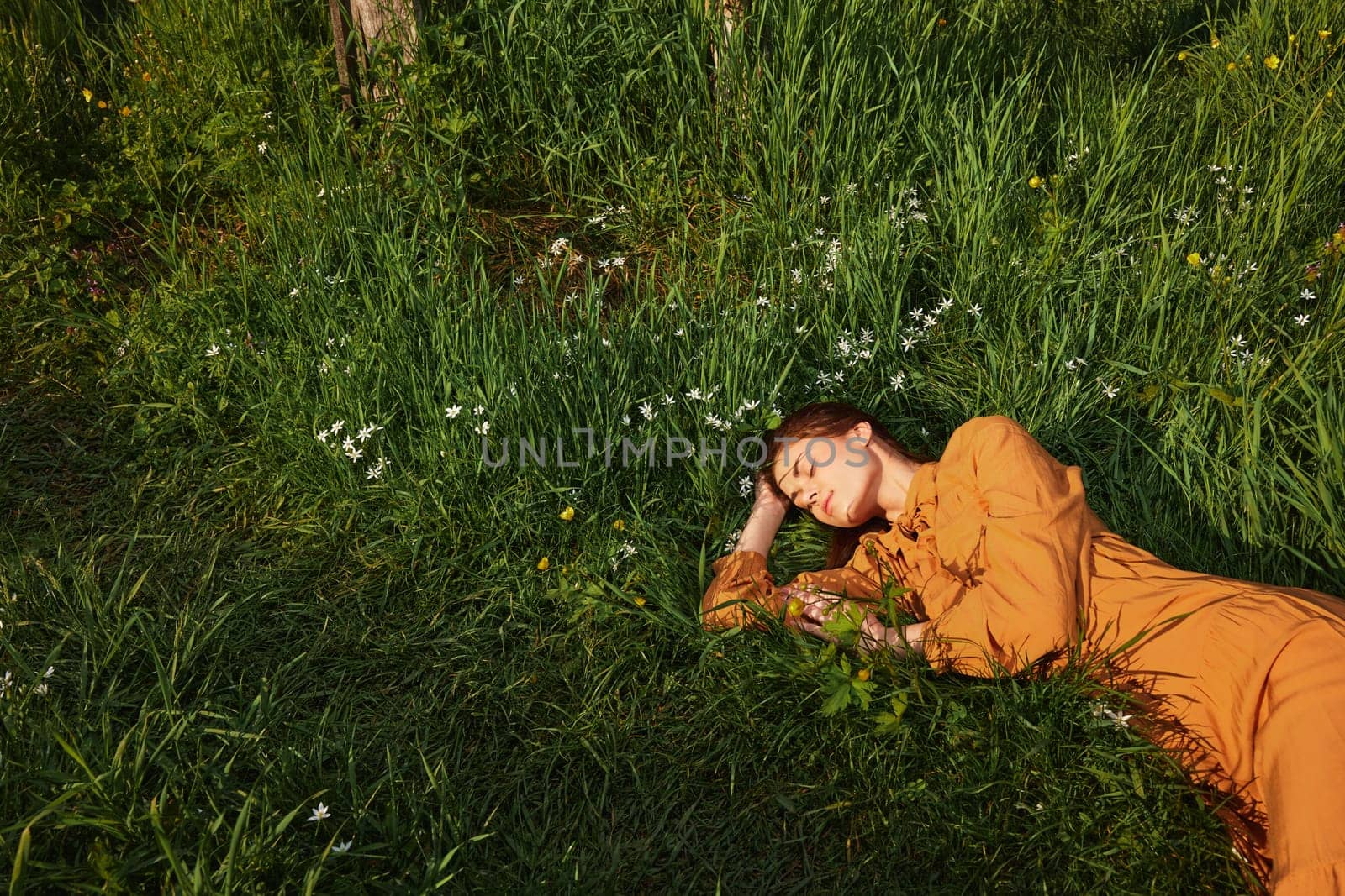 a calm woman with long red hair lies in a green field with yellow flowers, in an orange dress with her eyes closed, with a pleasant smile on her face, enjoying peace, illuminated by the setting sun by Vichizh