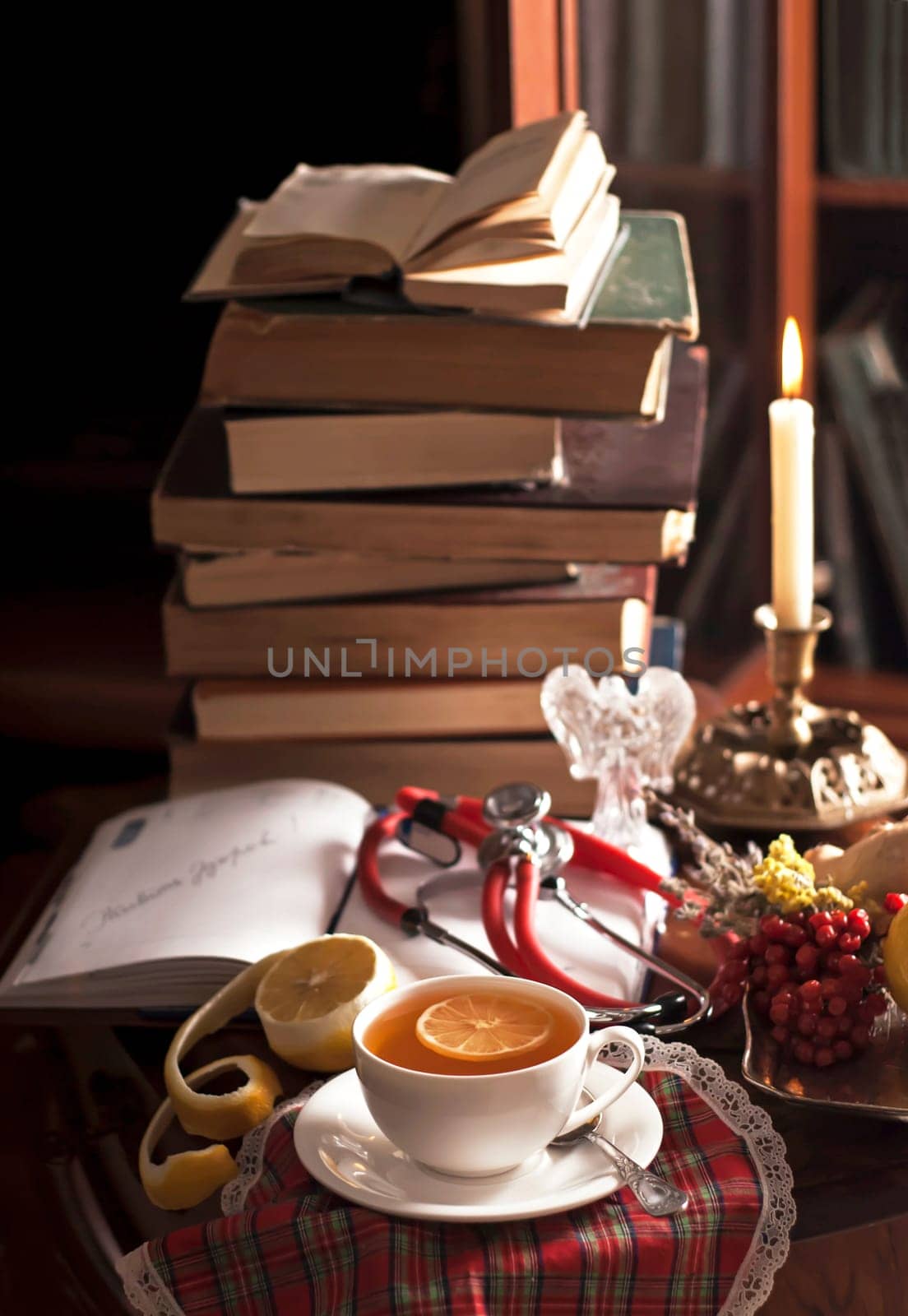 A stack of vintage books, a phonendoscope, a cup of tea with lemon, an open notepad, medicinal plants on a wooden table