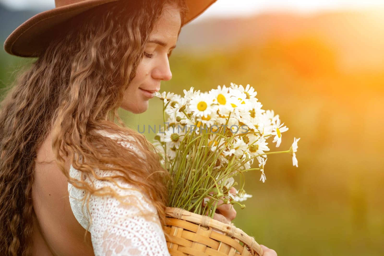 A middle-aged woman in a white dress and brown hat holds a large bouquet of daisies in her hands. Wildflowers for congratulations.