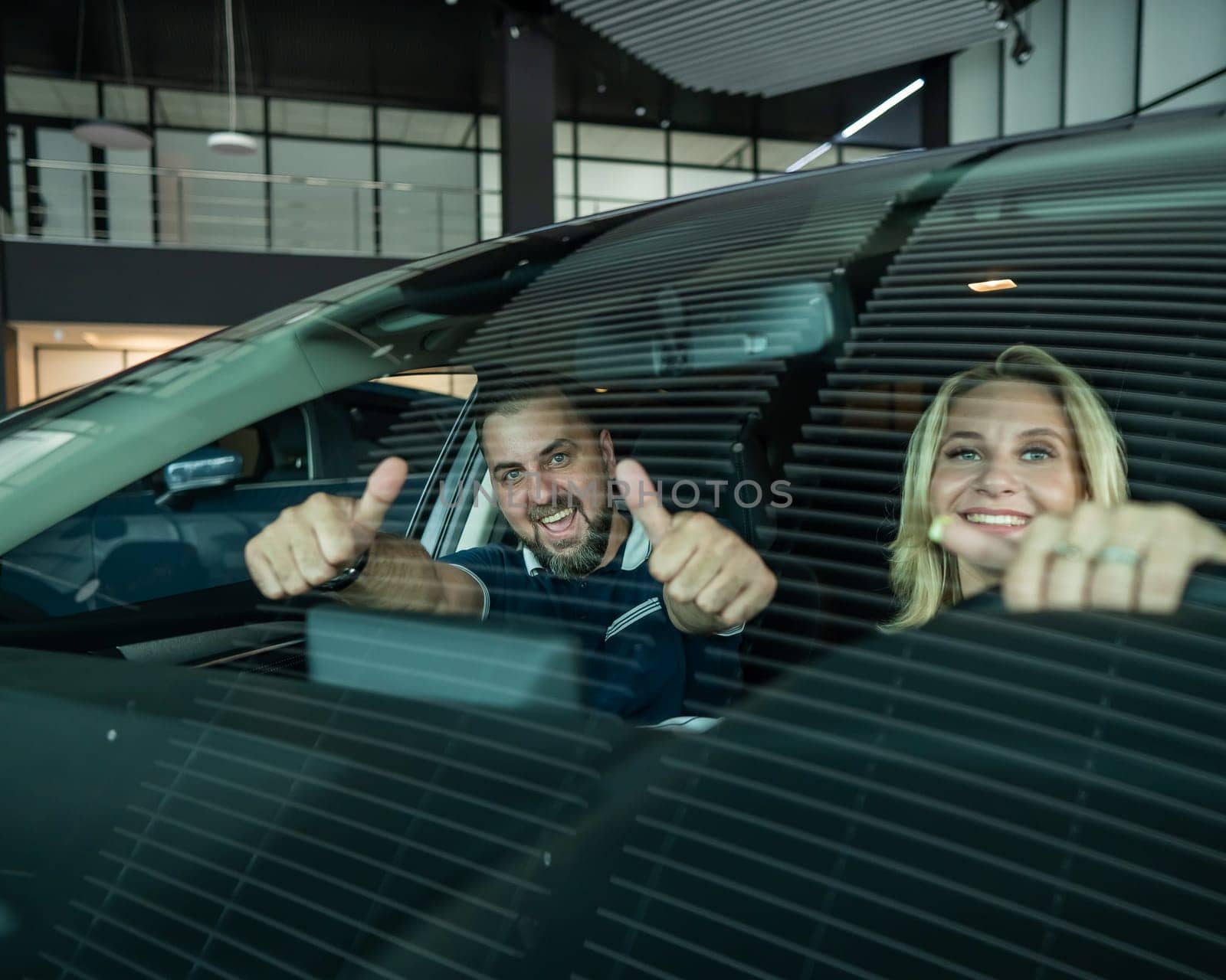 Happy caucasian couple is sitting in a new car in a car dealership. Man showing thumbs up. by mrwed54