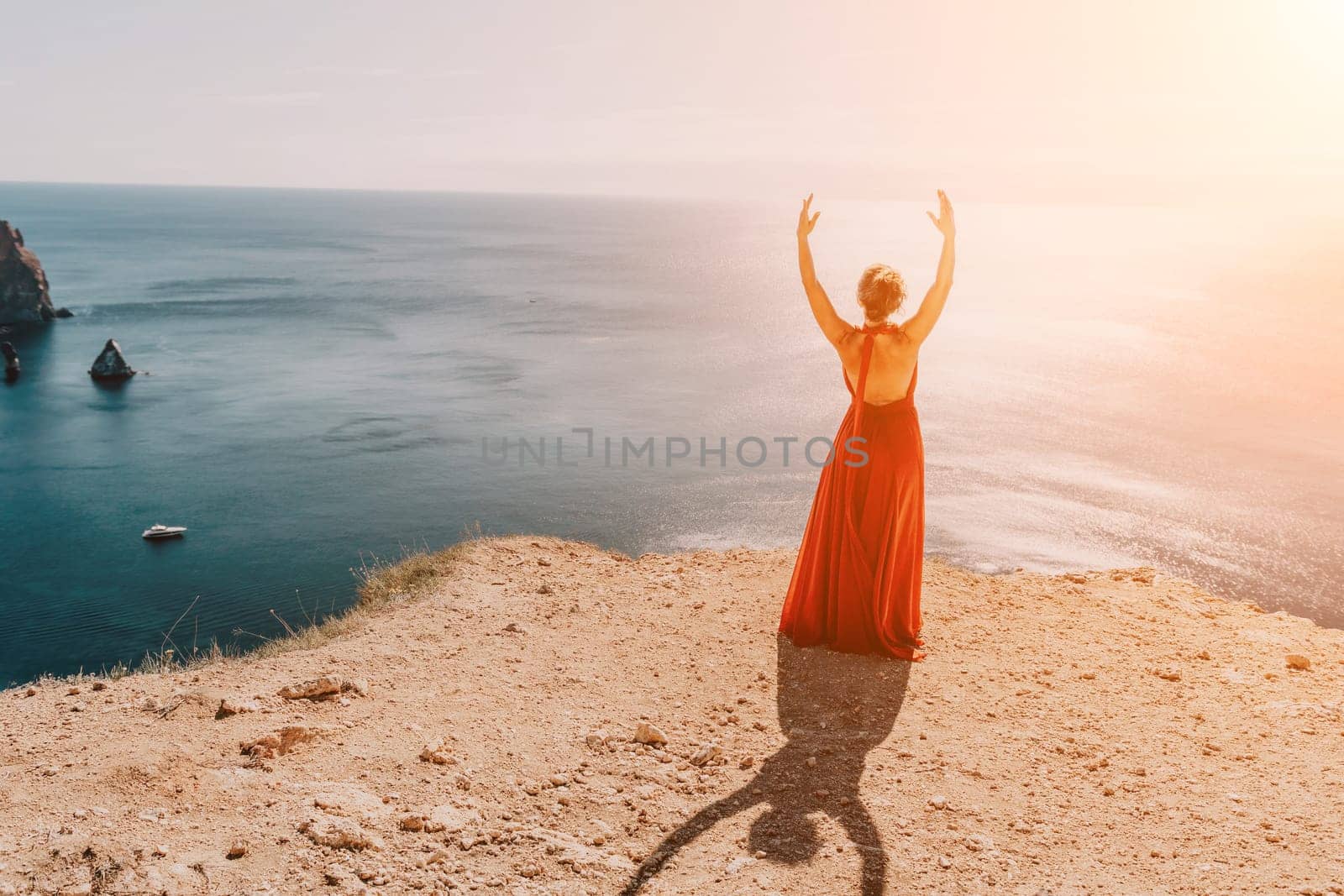 Side view a Young beautiful sensual woman in a red long dress posing on a rock high above the sea during sunrise. Girl on the nature on blue sky background. Fashion photo.