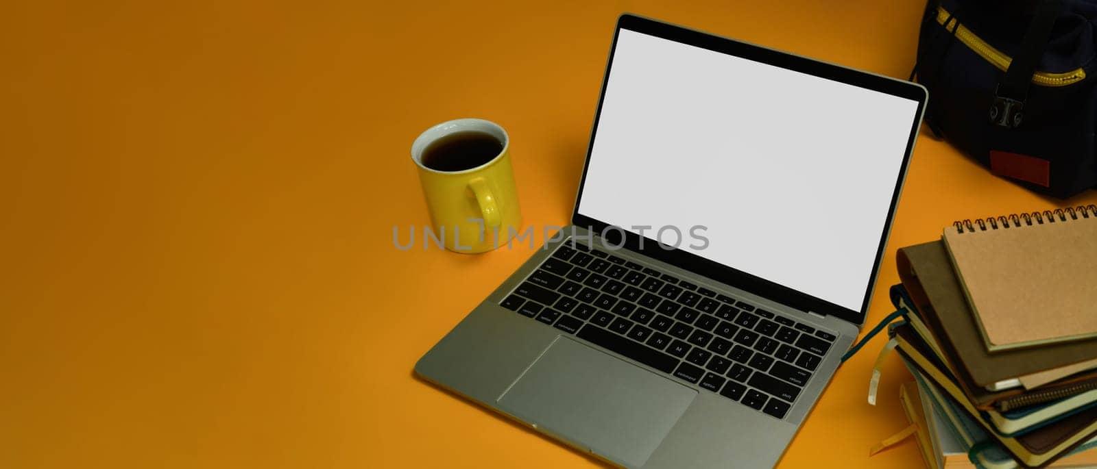 Laptop with blank screen, coffee cup, stack of books and backpack on yellow background. Online learning and education concept.