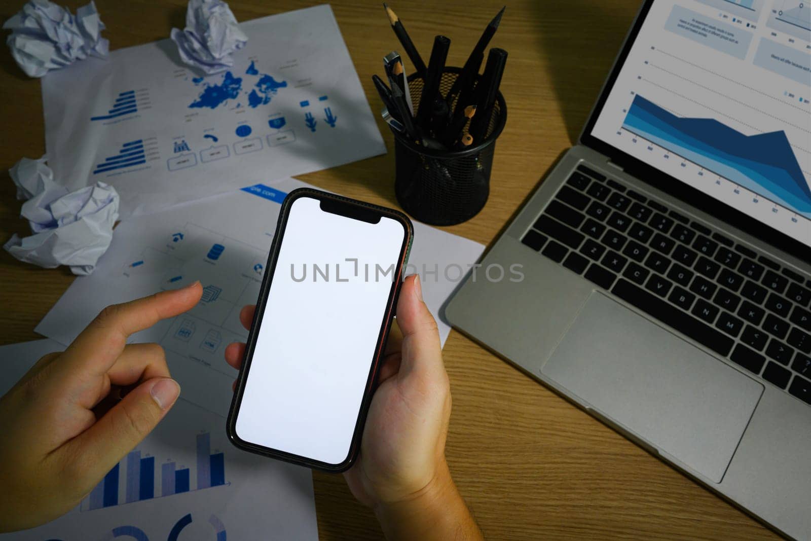 Hand holding smartphone, sitting in front of laptop at messy office desk. Close up view, blank screen for advertising text message.