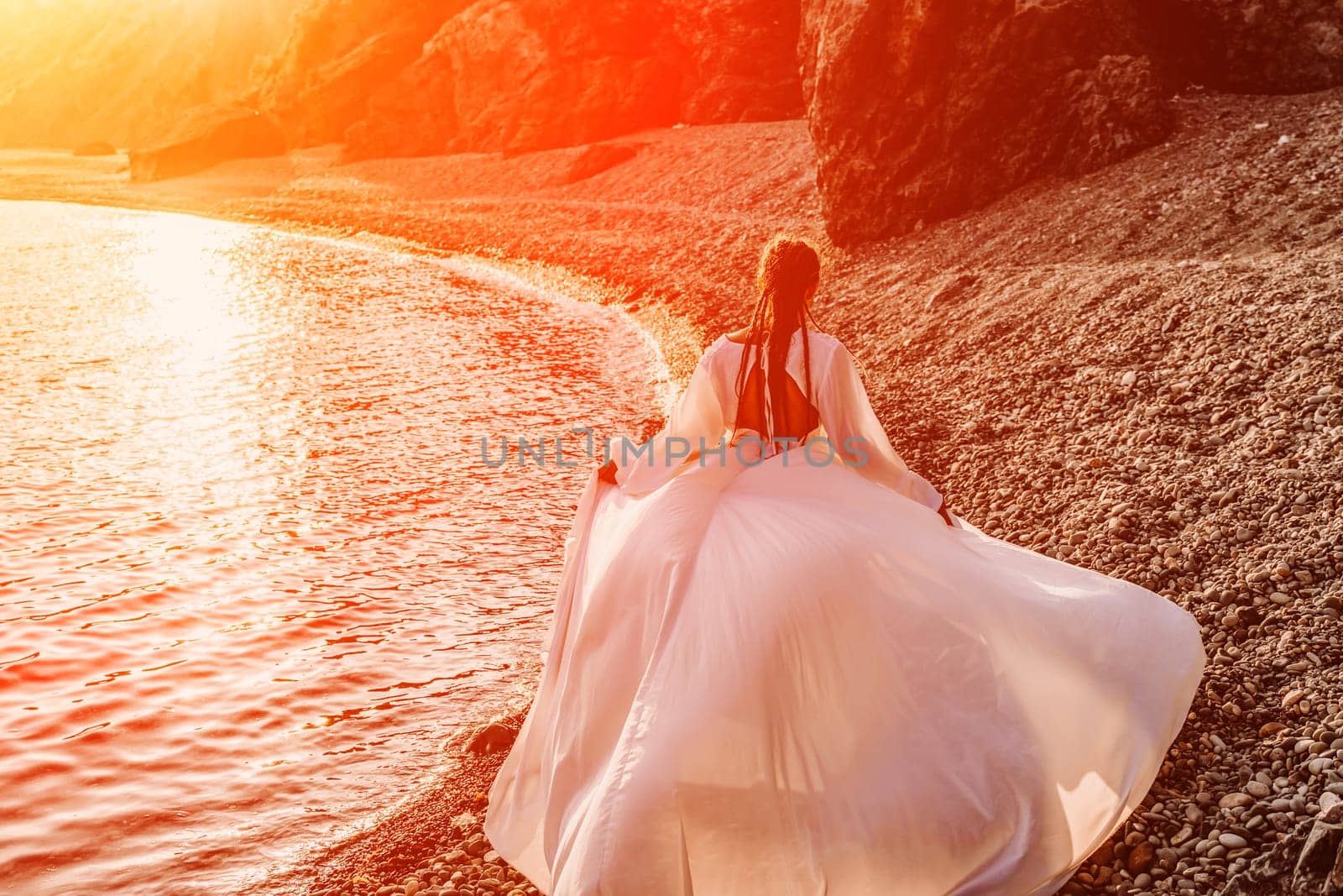 Mysterious woman silhouette long hair walks on the beach ocean water, sea nymph wind listens to the wave. Throws up a long white dress, a divine sunset. Artistic photo from the back without a face.