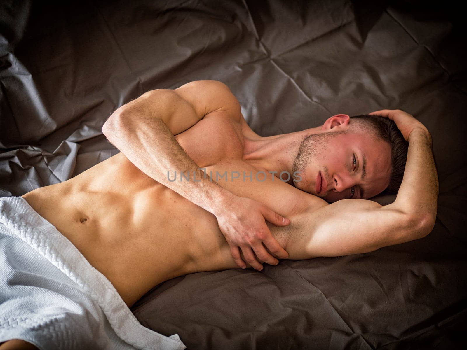 Shirtless sexy male model lying alone on his bed in his bedroom, looking at camera with a seductive attitude