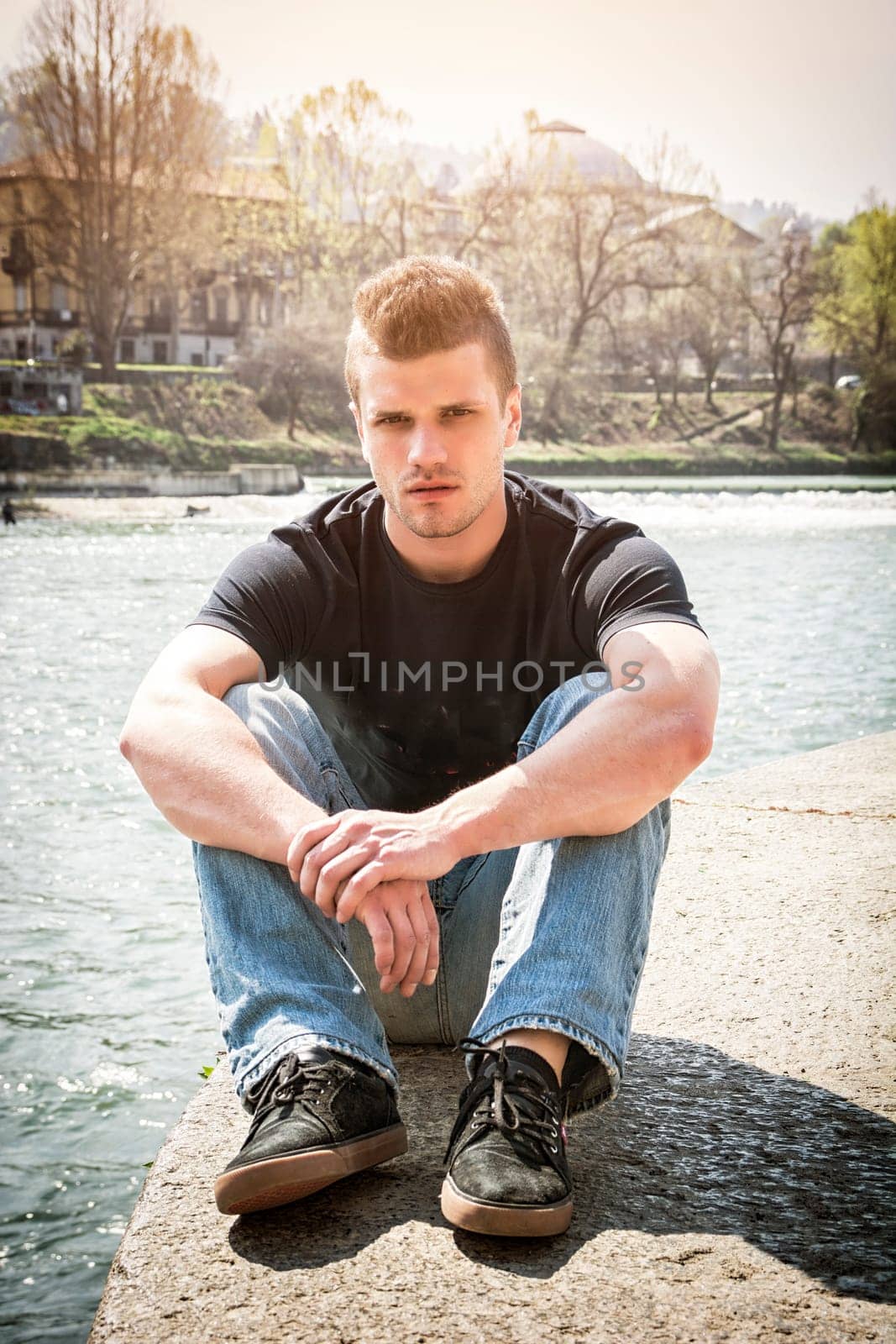 Full length of contemplative light brown haired young man wearing red hooded shirt and denim jeans sitting on wall beside picturesque river in Turin, Italy