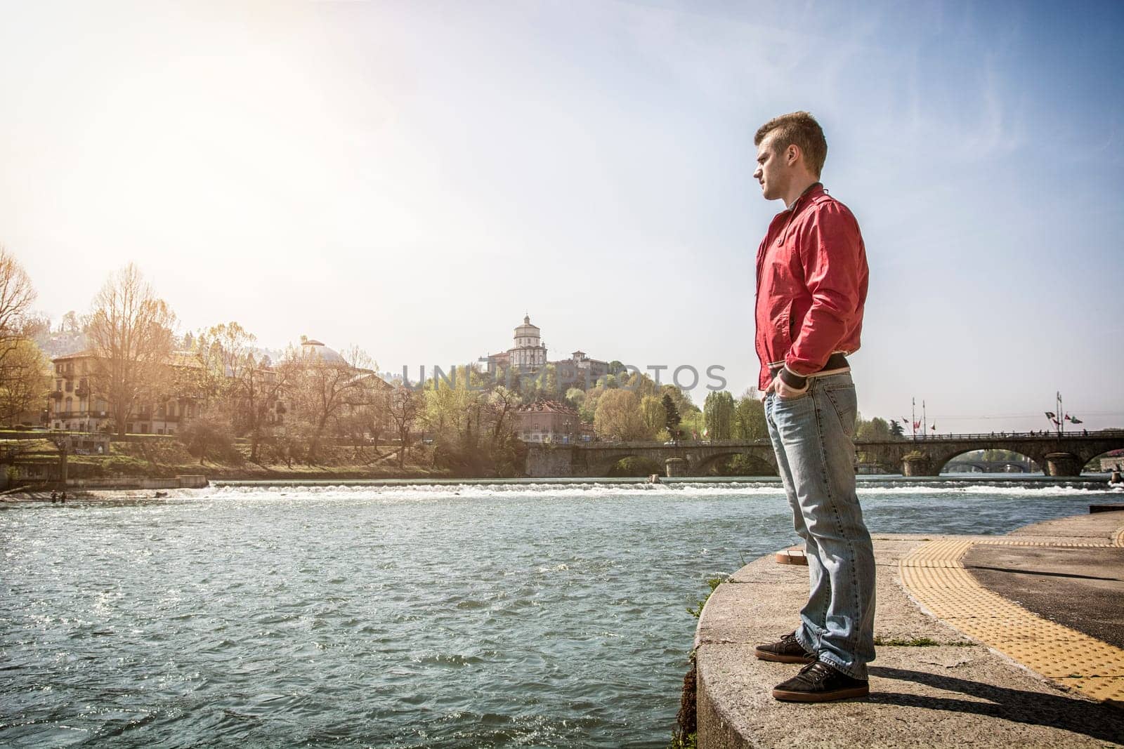 Full length of contemplative light brown haired young man wearing red hooded shirt and denim jeans standing beside picturesque river in Turin, Italy