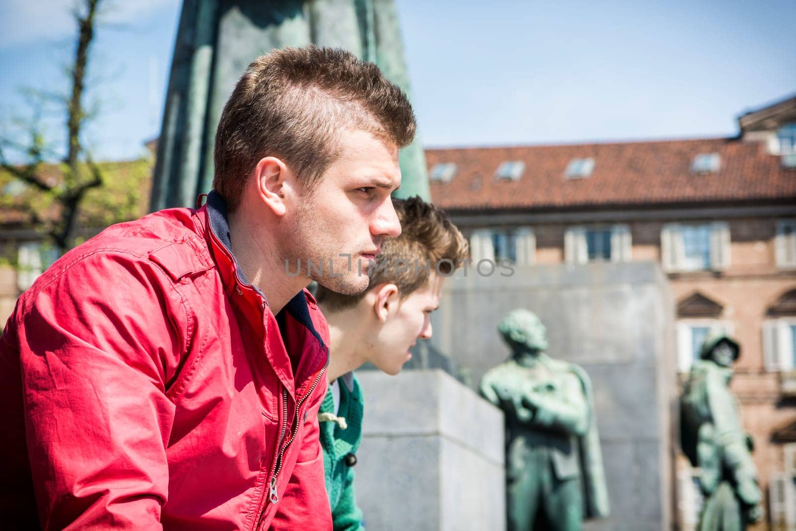 Two young men sitting on low wall in urban setting in European city. Summertime