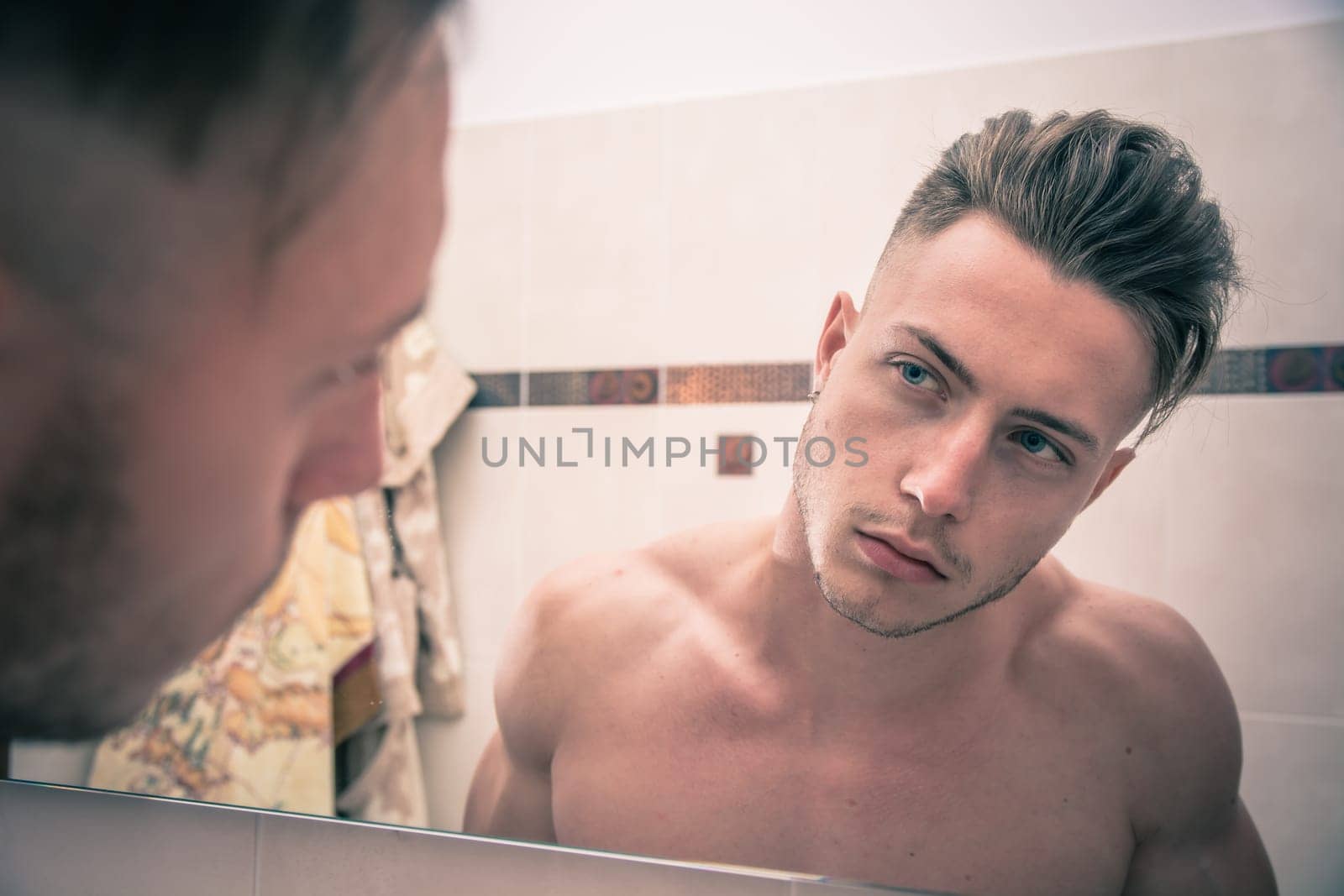 Shirtless handsome young man examining his stubble in bathroom mirror in the morning