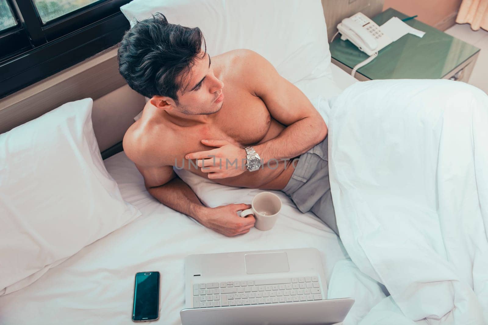 Naked young man with muscular body on bed with mug or cup in hand with coffee or tea, looking at laptop computer screen