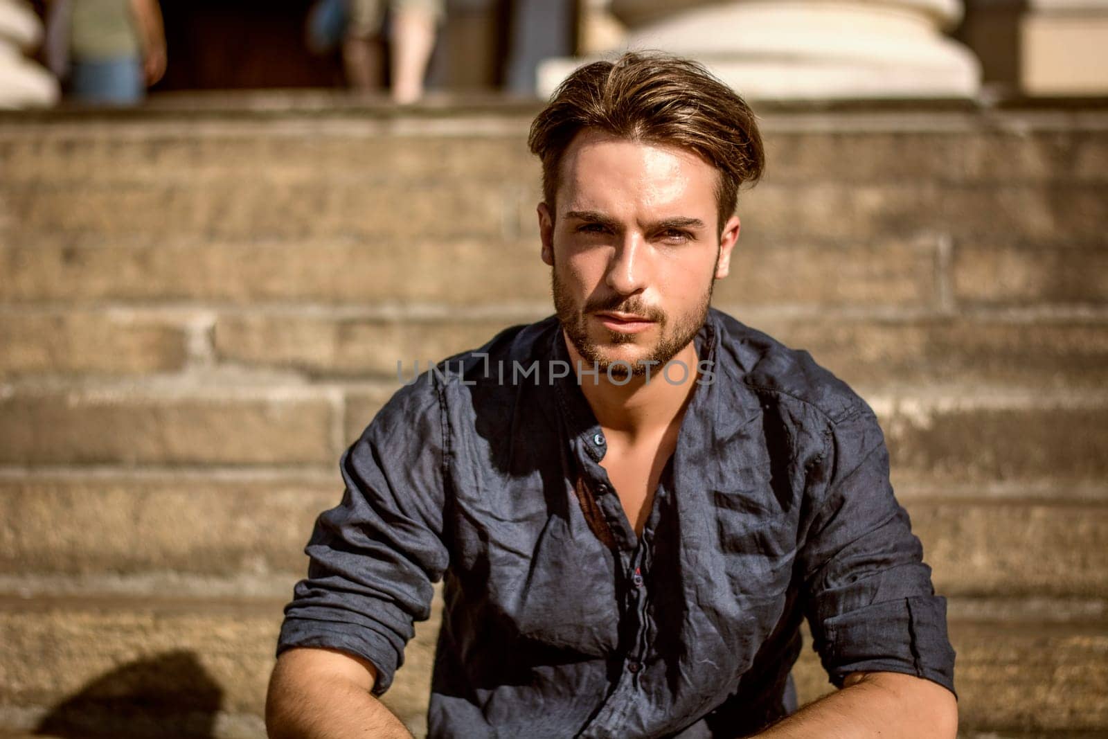 One handsome young man in urban setting in summer day, wearing blue shirt, sitting on stone steps