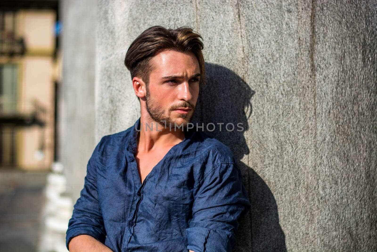 One handsome young man in urban setting in summer day, wearing blue shirt, leaning against stone column or pillar