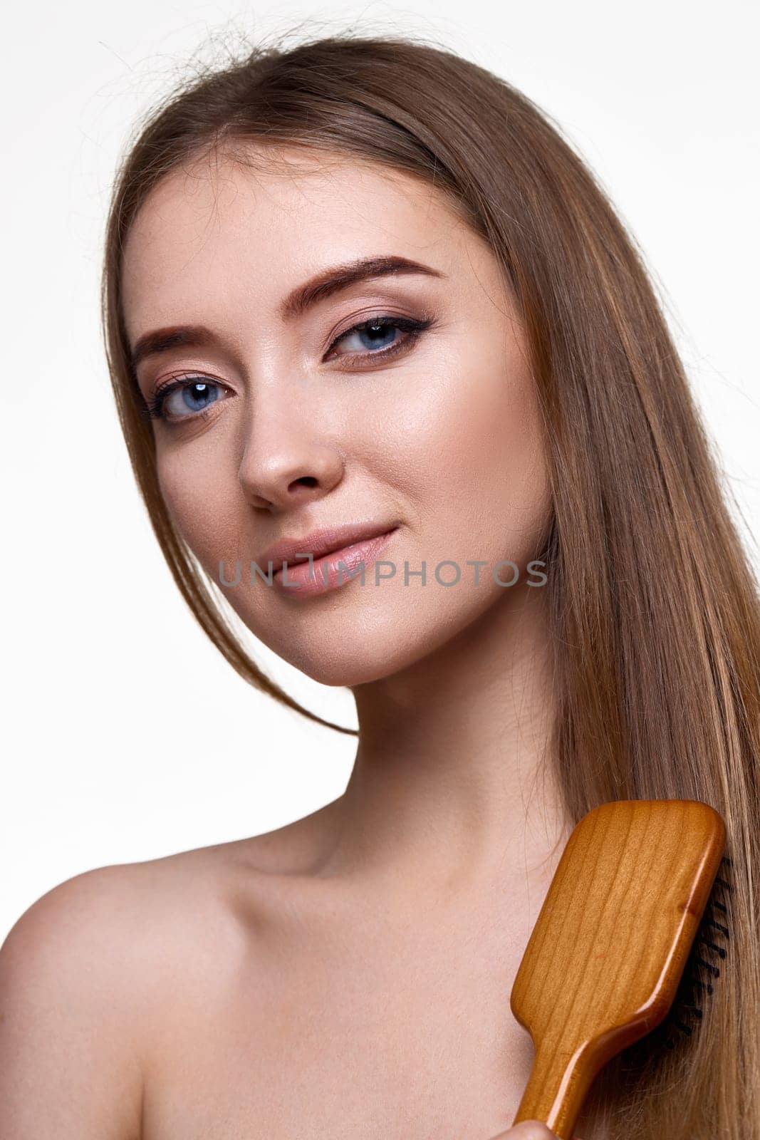 Beautiful smiling brunette woman looks at the camera and combs her hair with wooden brush isolated on white background