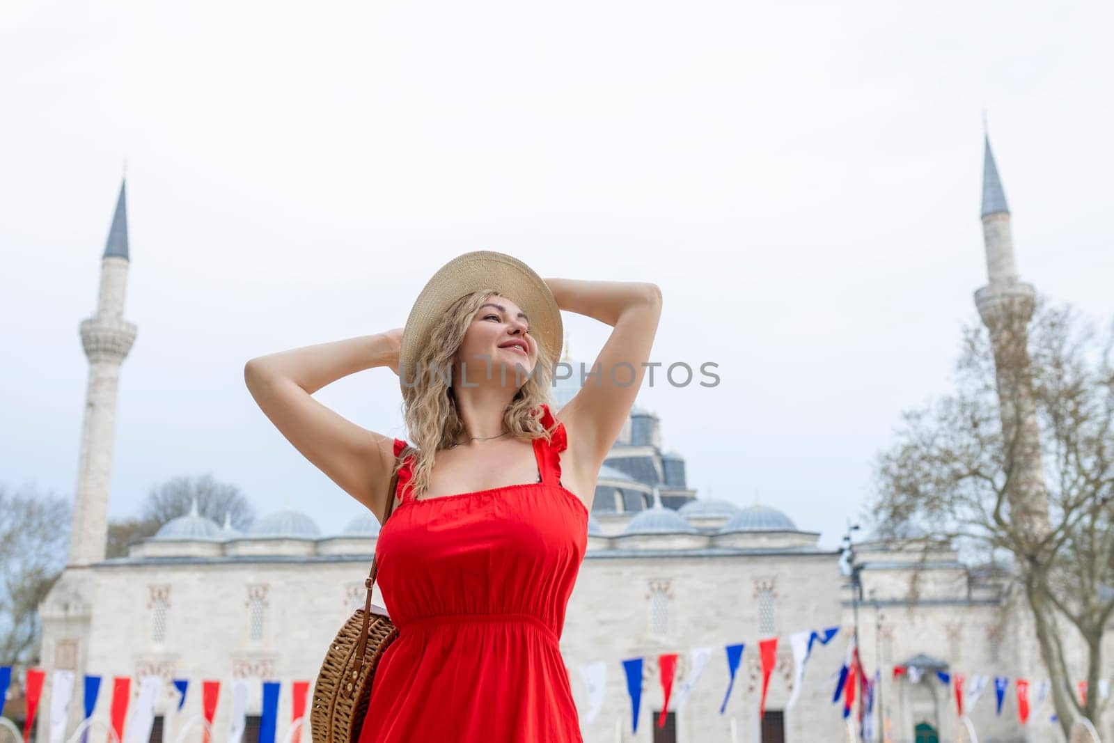 A beautiful traveler girl in a long red dress and a straw hat is photographed near the ancient sights of Istanbul in Turkey. Suleiman Blue Mosque in summer.