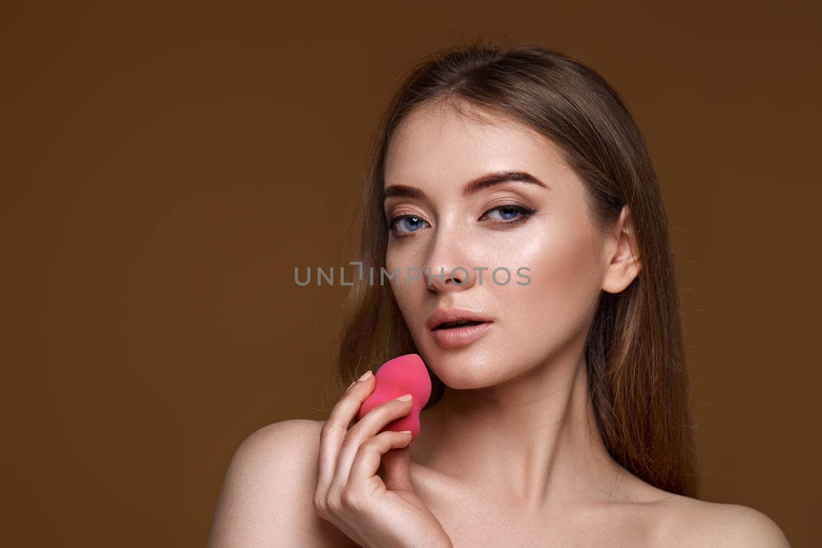 smiling young woman applying foundation on skin with beauty blender isolated on beige background