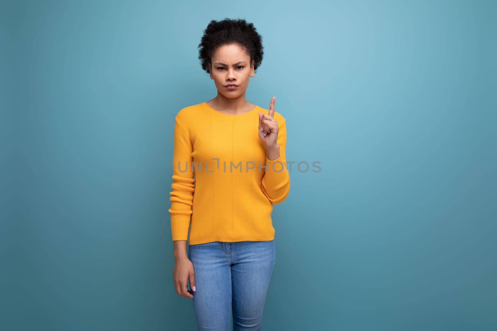 young Hispanic woman with black curly hair in a yellow sweater points her index finger up.