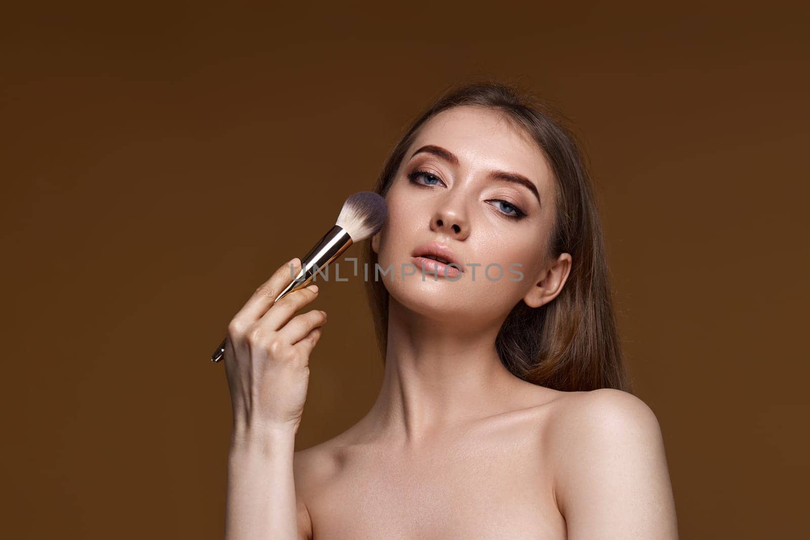 young woman applying blush on cheeks with make-up brush on beige background. copy space