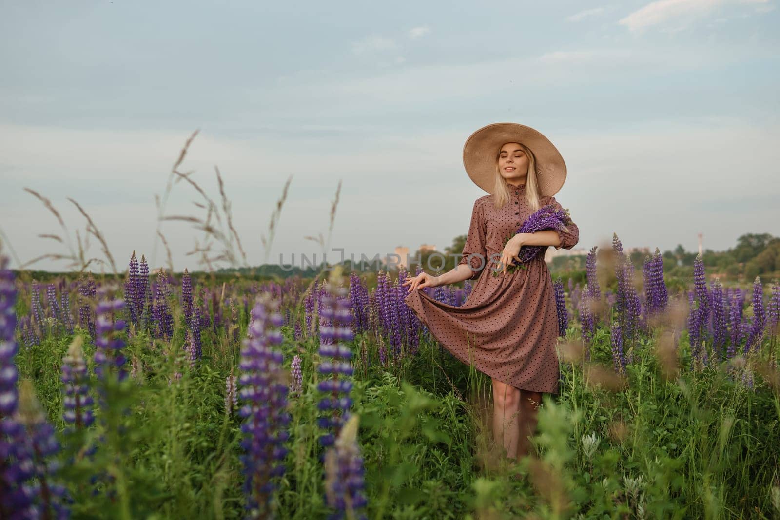 A beautiful woman in a straw hat walks in a field with purple flowers. A walk in nature in the lupin field.