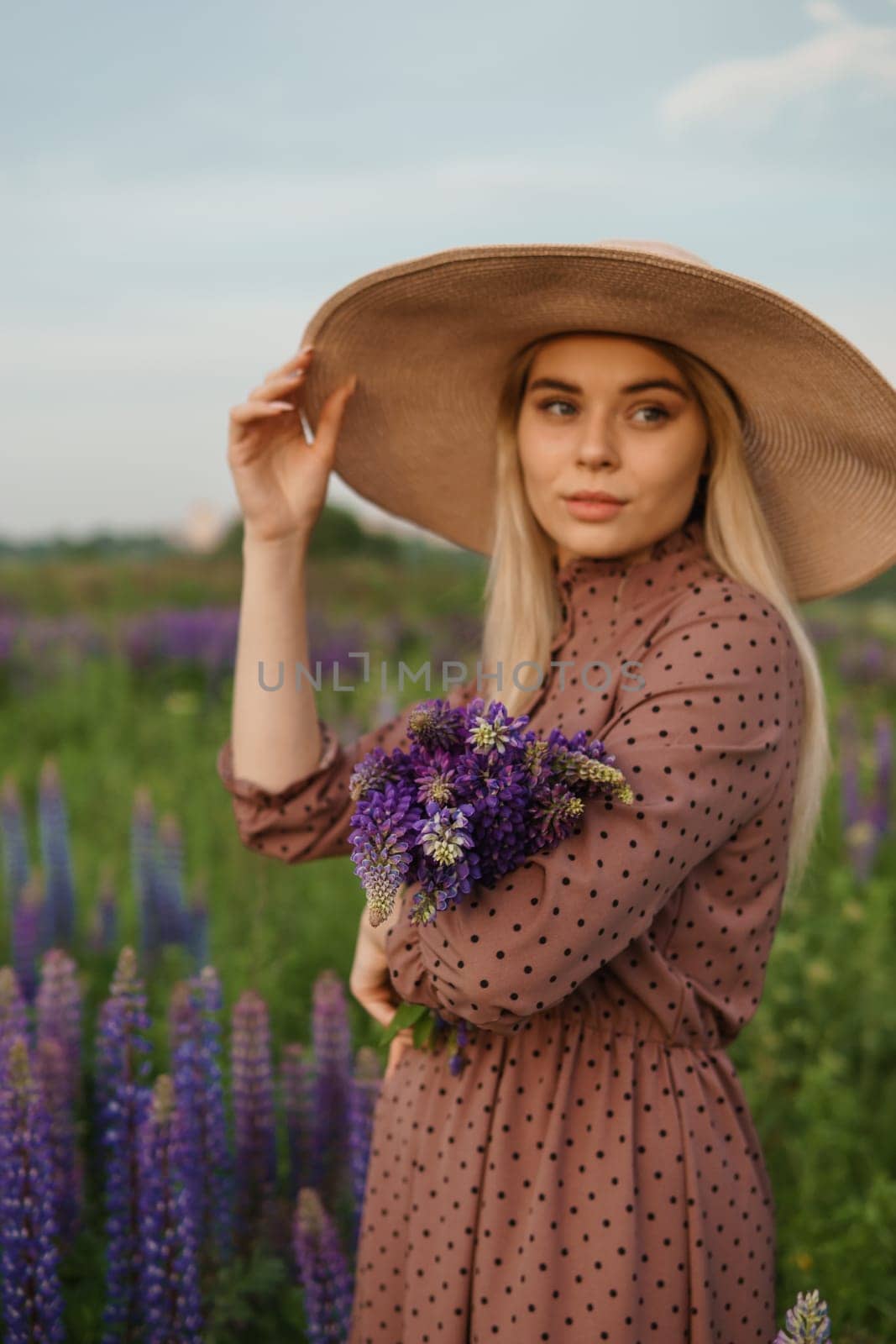 A beautiful woman in a straw hat walks in a field with purple flowers. A walk in nature in the lupin field.