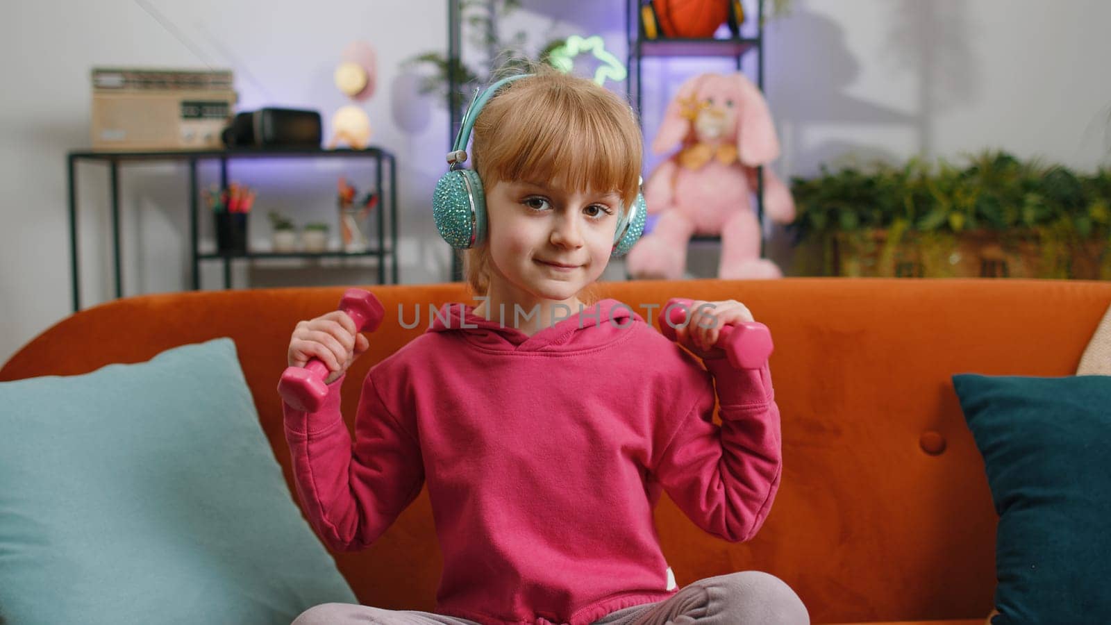 Young child kid girl lifting dumbbells while doing biceps exercises, enjoying training. Female teen toddler wearing wireless headphones listening music at home in room sits on sofa. Sport and fitness