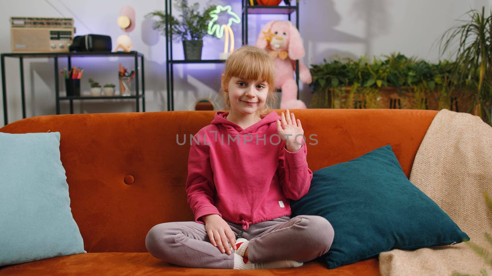 Young blonde child kid girl smiling friendly at camera and waving hands gesturing hello, hi, greeting or goodbye, welcoming with hospitable expression. Teen toddler at home in living room sits on sofa
