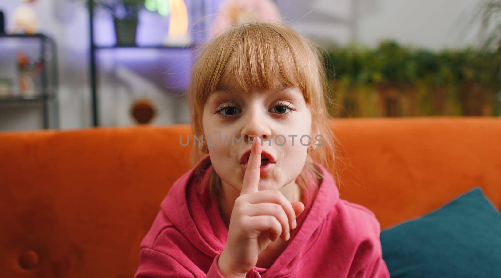 Shh be quiet please. Young blonde child kid girl presses index finger to lips makes silence hush gesture sign do not tell gossip privacy secret. Female teen toddler at home in living room sits on sofa