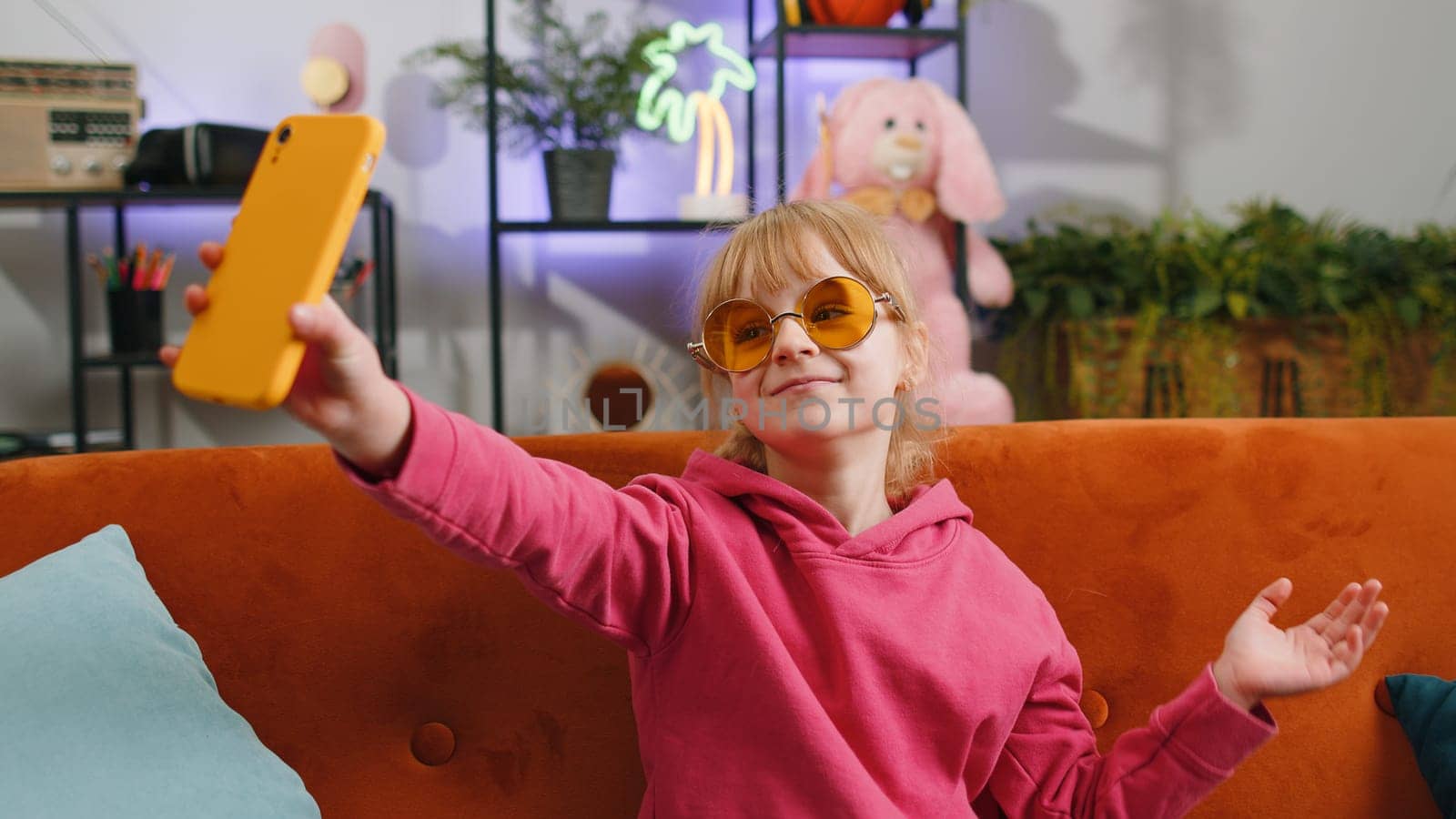 Young blonde child kid girl blogger taking selfie on smartphone, communicating video call online with social media subscribers. Female teen toddler recording video story at home in room sits on sofa