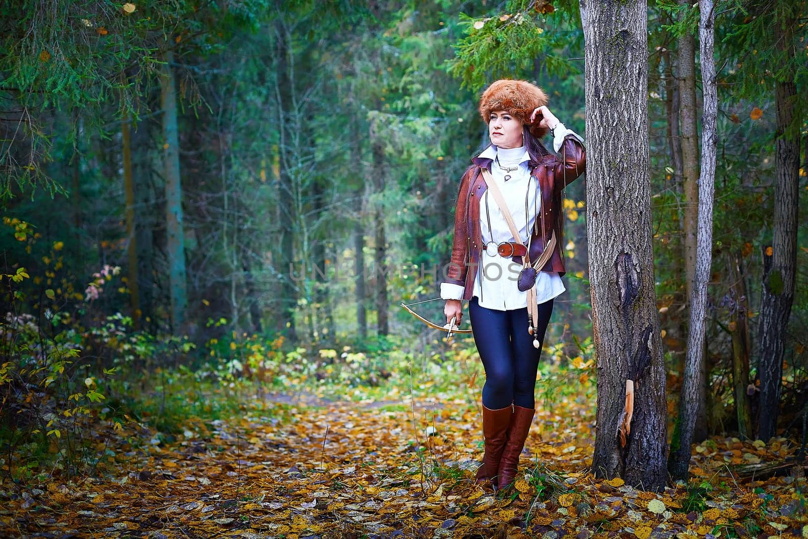 Girl in a leather jacket, a big red fox fur hat and with a crossbow in the forest in autumn. A female model poses as a fabulous royal huntress on nature hunt at a photo shoot