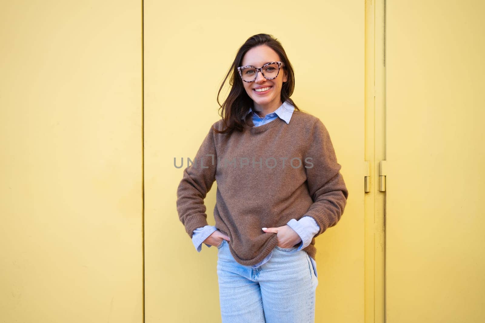 Happy woman in glasses outdoor on yellow color background. Positive people concept. Smiling girl looking at camera, hands in pocket, dressed sweater and jeans