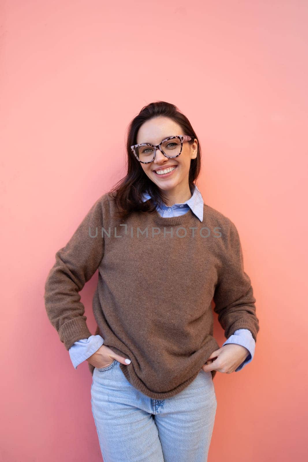 Happy woman in glasses outdoor on yellow and pink color background. Positive people concept. Smiling girl looking at camera, hands in pocket, dressed sweater and jeans