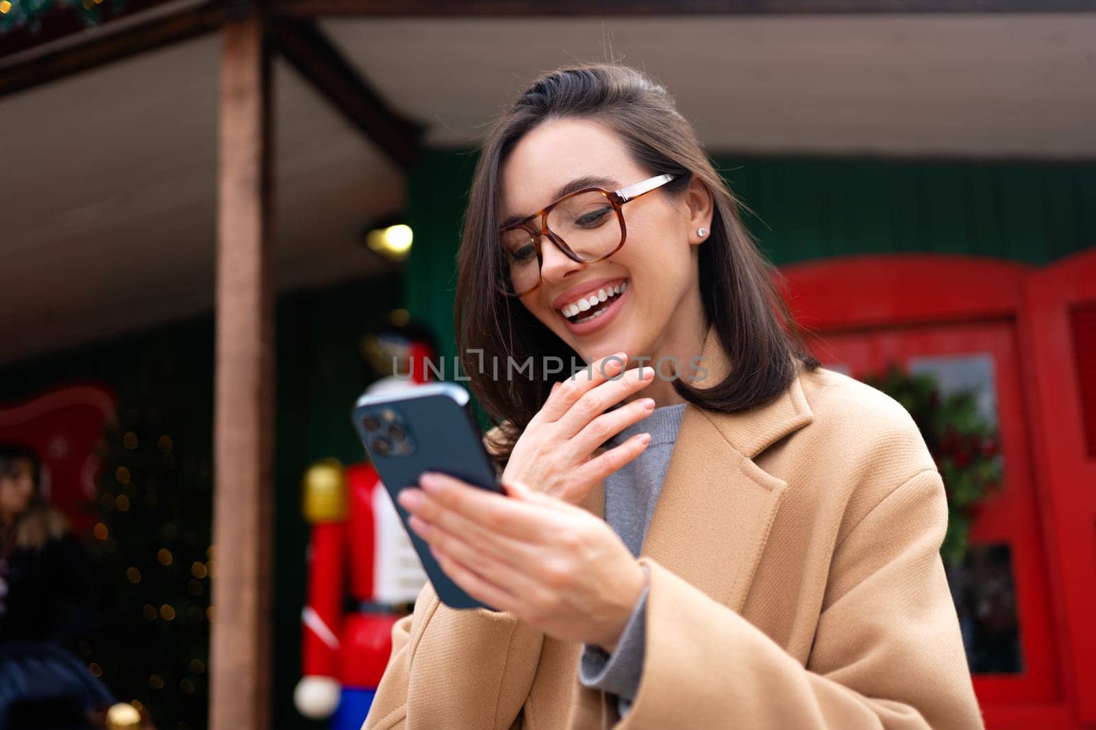 Young beautiful woman in glasses using mobile phone at day in the city. Happy woman using smartphone to do a video call on a winter day while looking at camera.