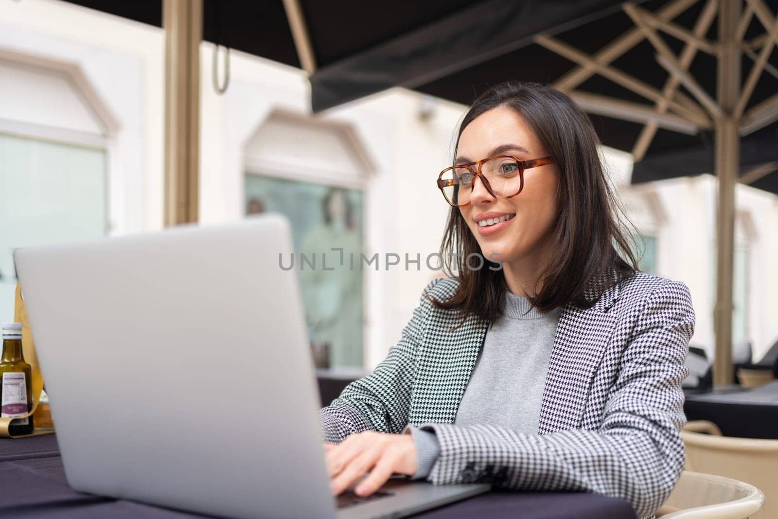 Woman working on remote from outdoor cafe, sitting with laptop on restaurant terrace on city street, studying. Female freelancer using laptop, smiling looking laptop screen