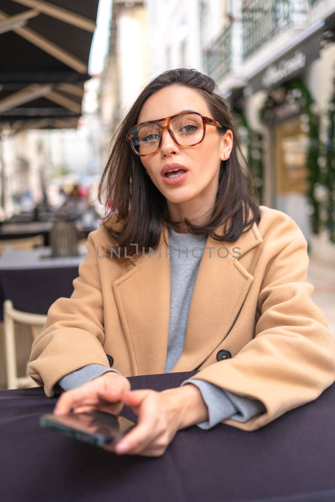 Middle-aged woman using smartphone sitting outdoor cafe. Confidence businesswoman in glasses dressed trench coat holding smartphone in hand looking screen