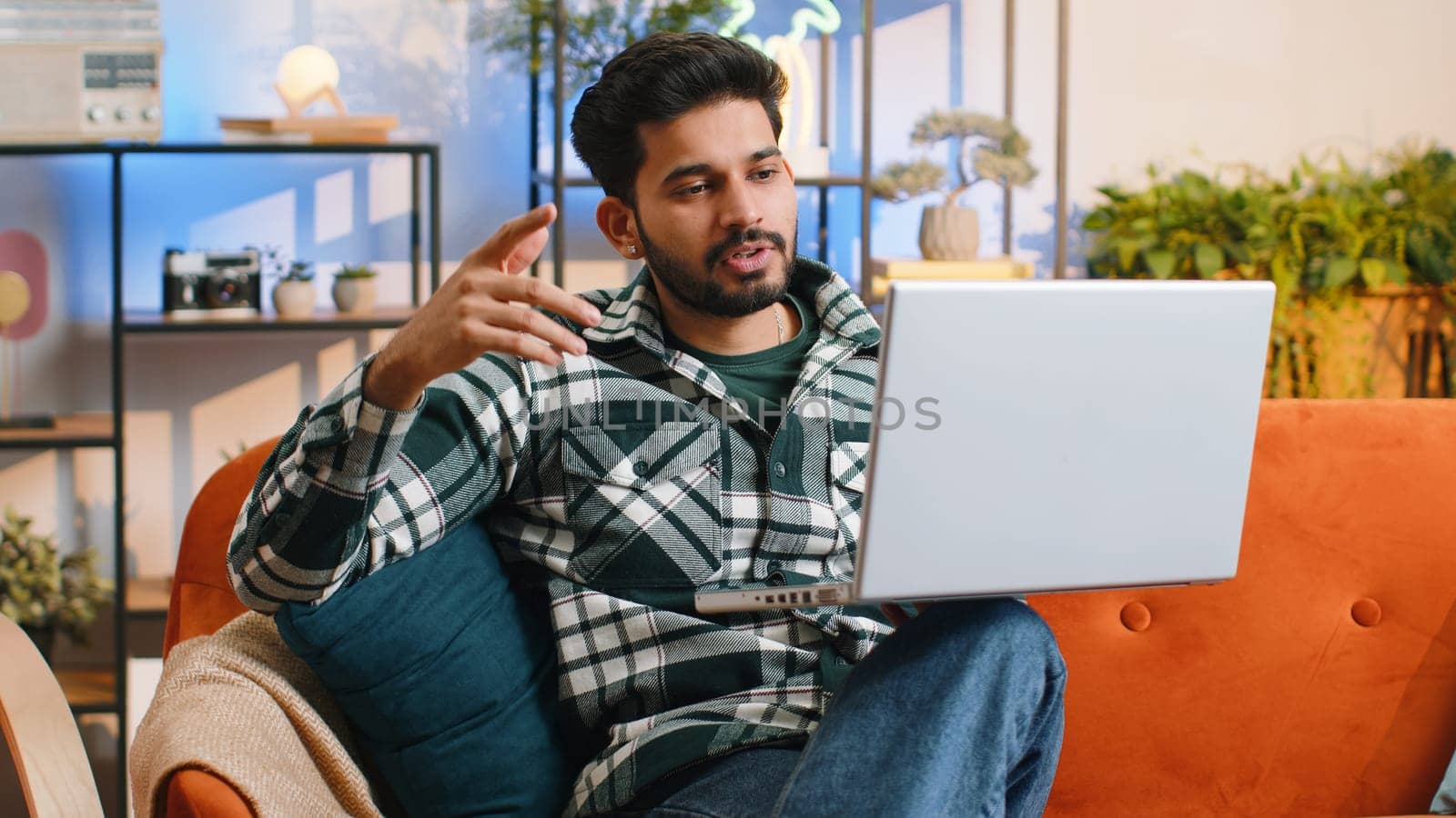Indian man sitting on couch looking at camera, making video webcam conference call with friends or family, enjoying pleasant conversation. Young hindu guy laughing waving hello at home room
