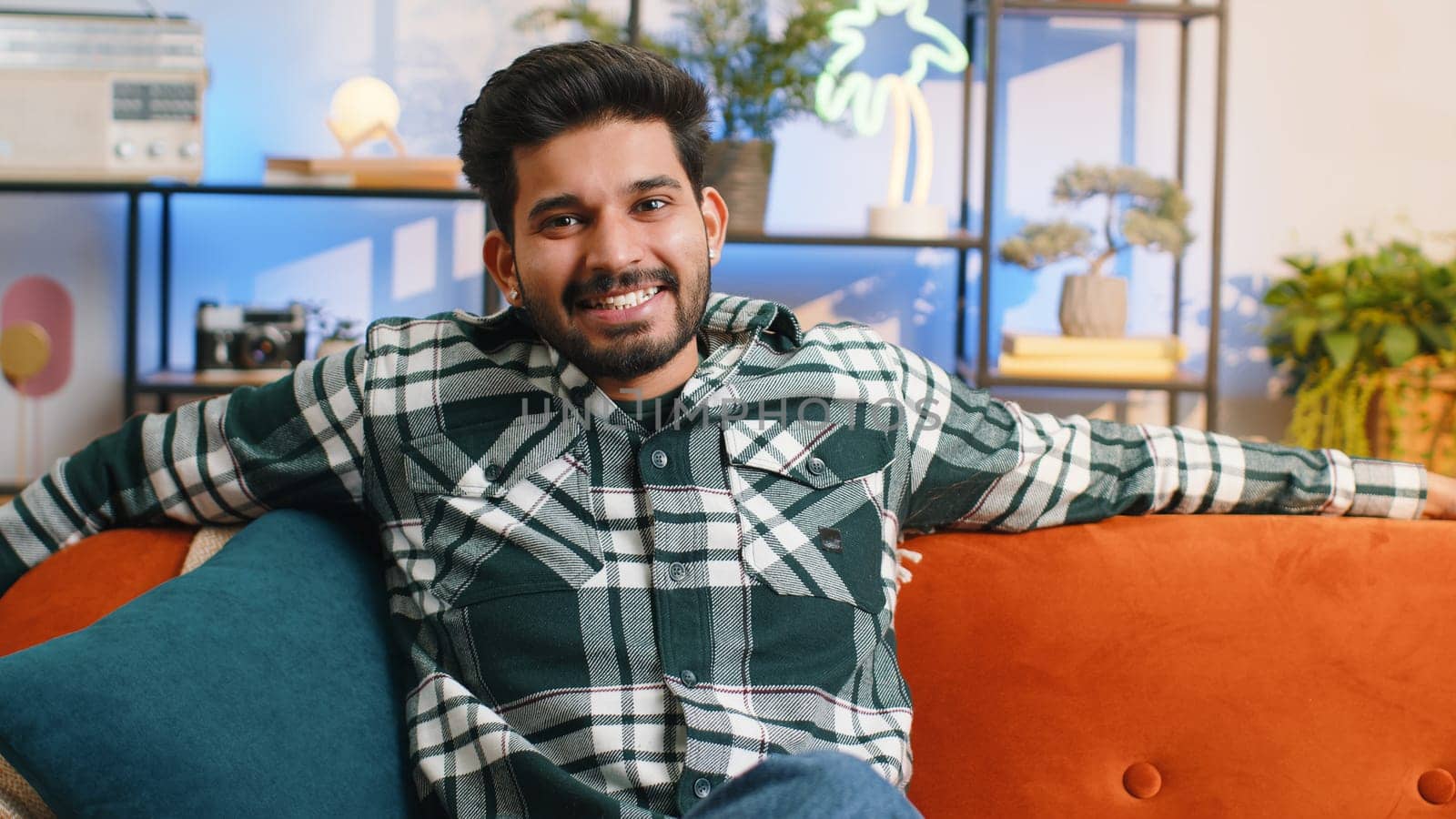 Portrait of happy calm indian man at home couch smiling friendly, glad expression looking away dreaming resting, relaxation feel satisfied concept good news, celebrate win. Hindu guy in living room