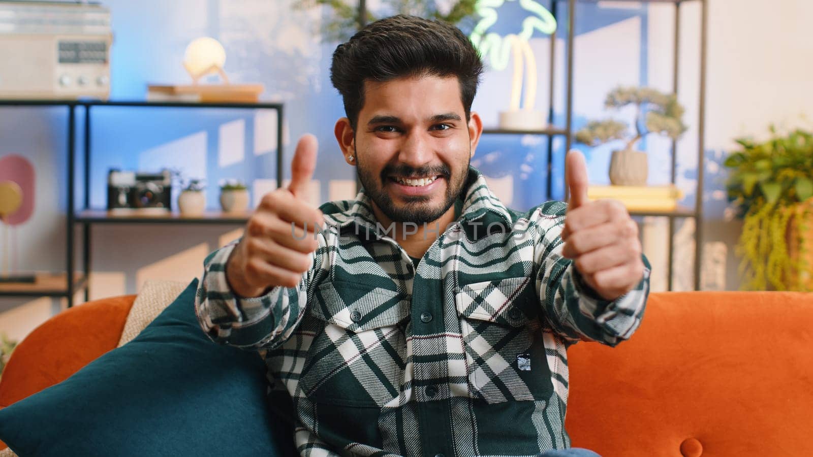 Like. Happy excited indian man looking approvingly at camera showing thumbs up, like sign positive something, good great news, positive feedback. Young hindu guy sitting on couch at home living room