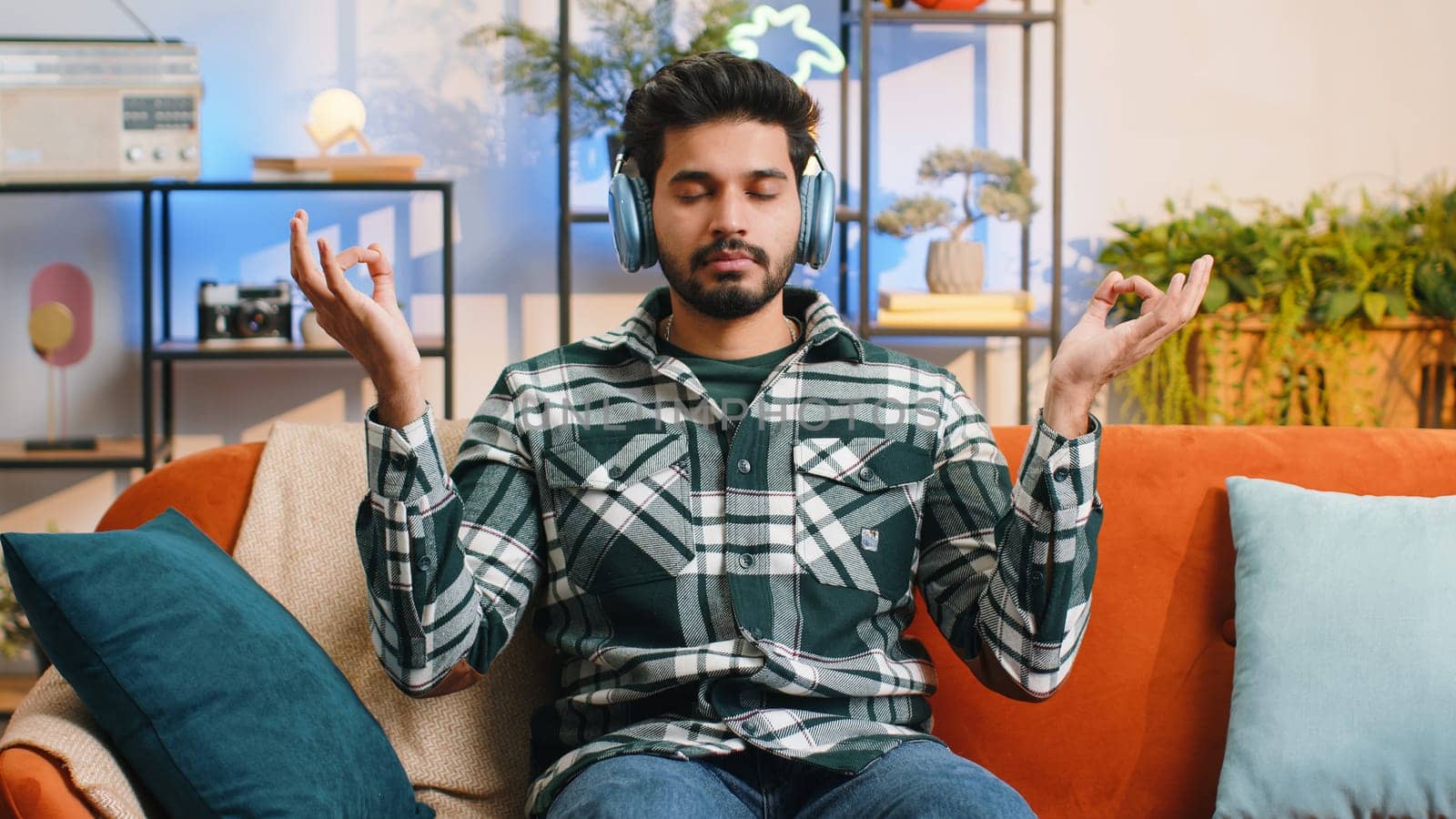 Keep calm down, relax, inner balance. Indian man breathes deeply with mudra gesture, eyes closed, meditating with concentrated thoughts, peaceful mind at home apartment. Hindu guy sitting on couch