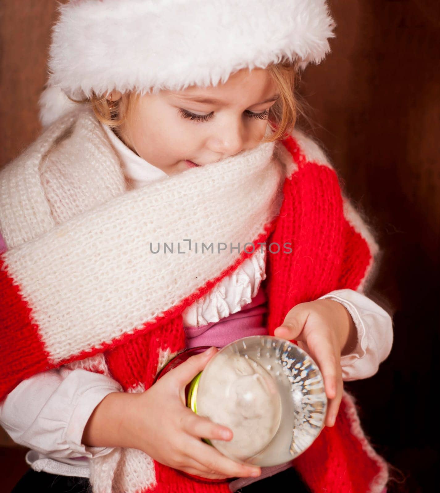 A girl in a warm red scarf and a santa claus hat examines a toy big snow globe. The child rejoices in winter. New Year cartoons and the holiday of Christmas by aprilphoto