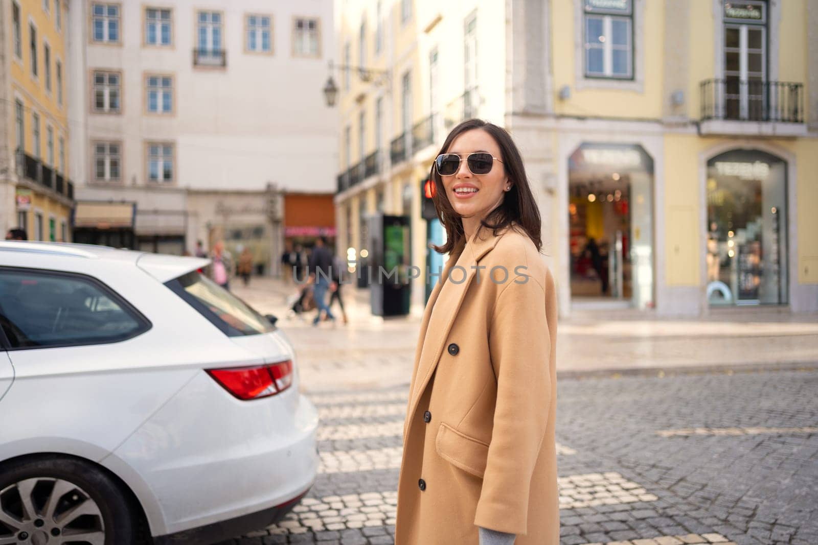 Happy caucasian girl walking city street on sunny autumn day. Brunette woman wears stylish trench coat and sunglasses. Positive emotions lifestyle concept