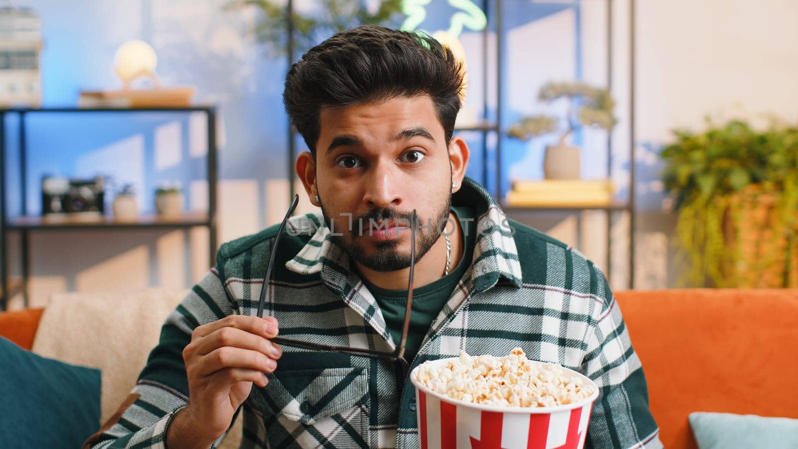 Excited indian man in 3D glasses sits on sofa eating popcorn snacks and watching interesting TV serial sport game, film, online social media movie content at home apartment. Guy enjoying entertainment
