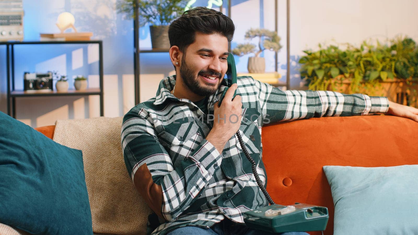 Portrait of indian man making wired telephone conversation with friends sitting call on couch at home in room. Happy excited guy enjoying old-fashioned retro vintage phone from 90s talking indoors