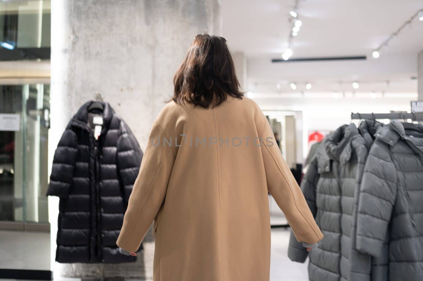 Rear view unrecognizable woman walking through clothing store watching winter collection chooses what to buy dressed stylish trench coat