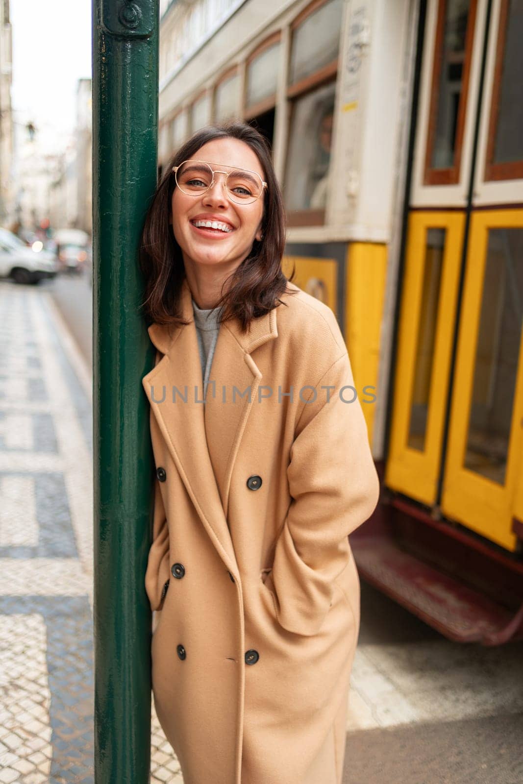 Happy caucasian girl walking city street on sunny autumn day. Brunette woman wears stylish trench coat and glasses. Positive emotions lifestyle concept. Vertical photo