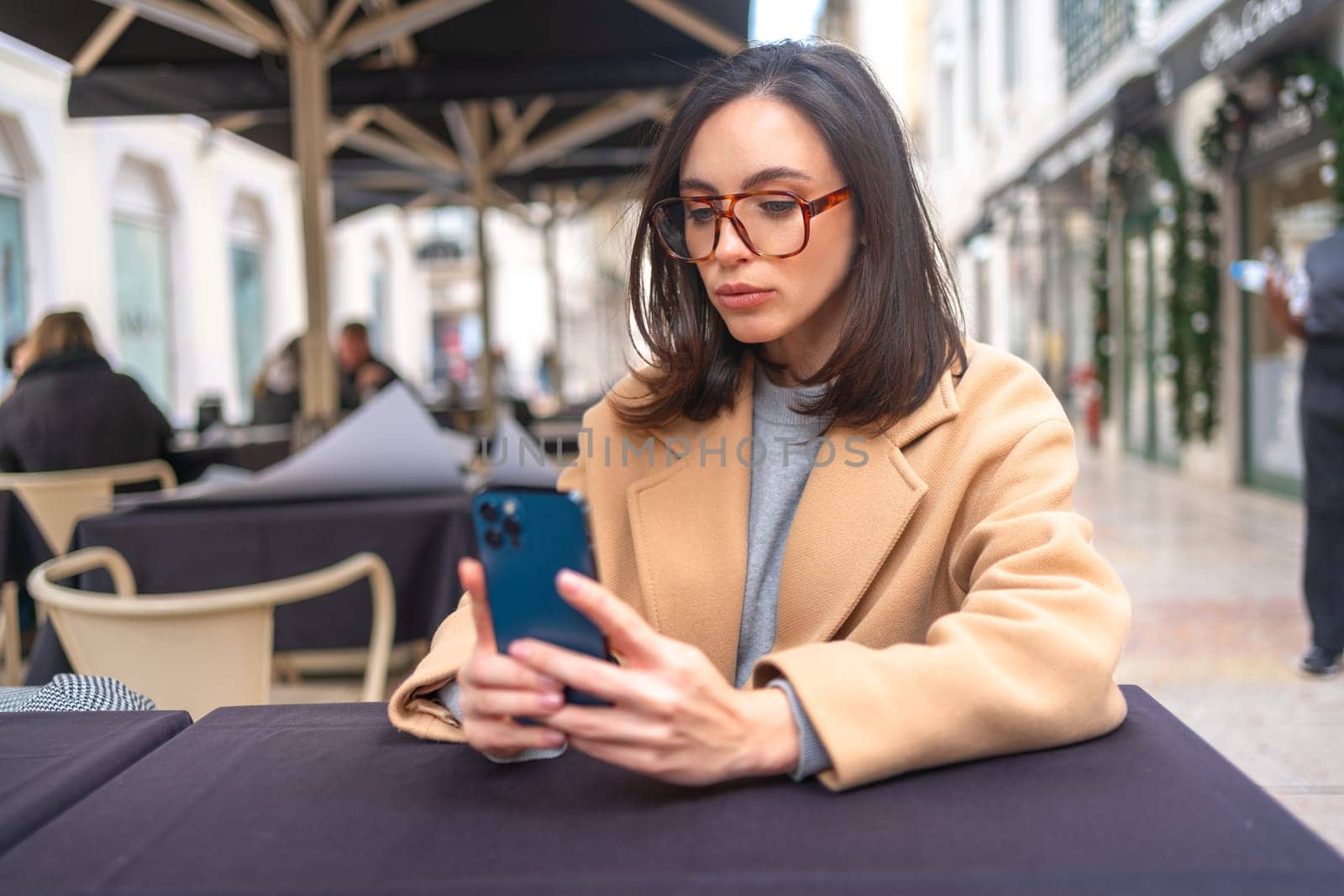 Serious middle-aged woman using smartphone sitting outdoor cafe. Focused businesswoman in glasses dressed trench coat holding smartphone in hand looking screen