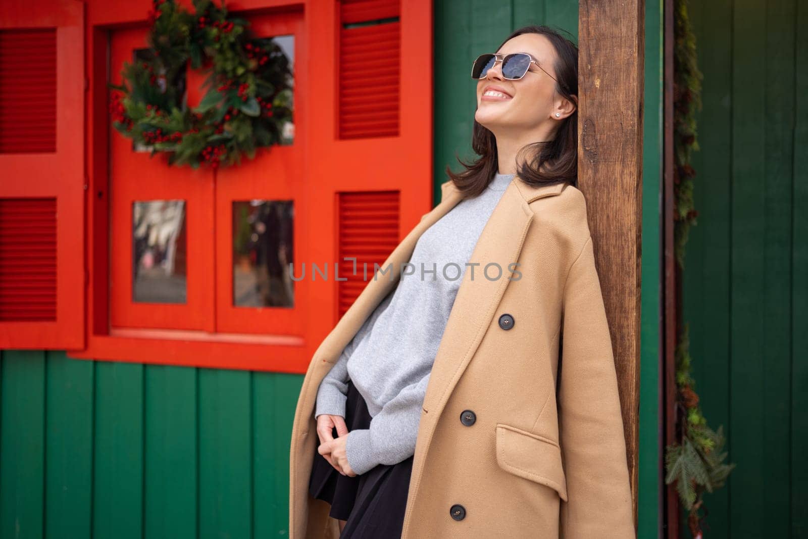 People, season and Christmas concept portrait of happy smiling middle age woman outdoors dressed stylish cream trench coat with Christmas festive decoration on background.